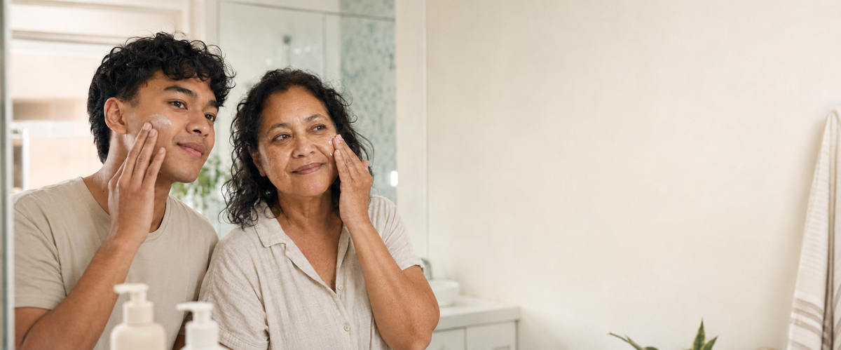 A young man and an older woman apply skincare creams to their faces while looking into a bathroom mirror. Bottles of skincare products are visible on the counter in the foreground.